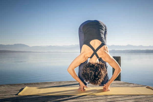 Vrouw die yoga beoefent op een oefenmat op de steiger boven het meer tegen een heldere lucht