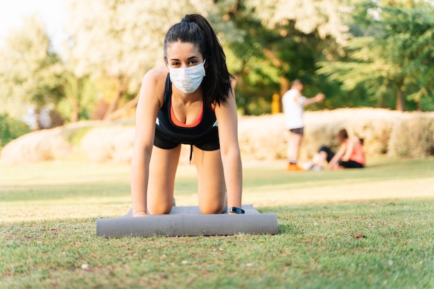 Vrouw die yoga met een matras in een park doet dat een masker draagt. Nieuw normaliteitsconcept