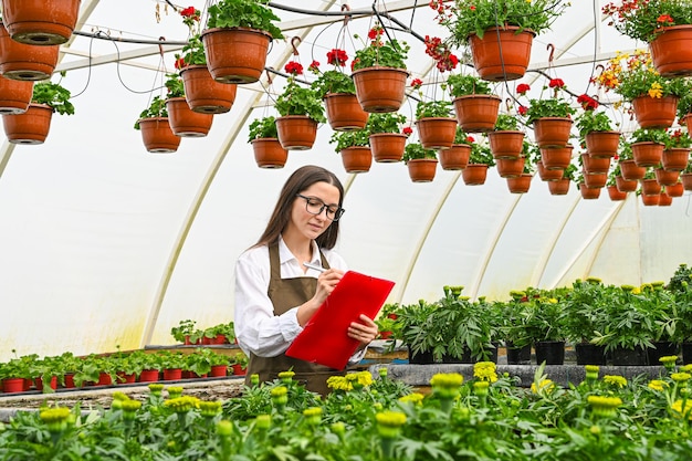 Vrouw doet lijst met planten Portret van een jonge vrouw aan het werk in kas in uniform en klembord in haar hand