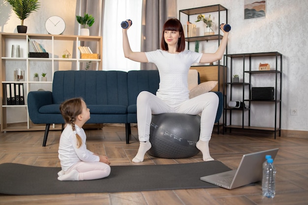 Vrouw doet sport zittend op fitball met halters in handen terwijl haar dochter op de mat zit