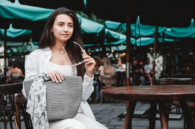 Vrouw draagt een zonnebril en witte kleren met een gebreide tas aan de tafel van een straatcafé