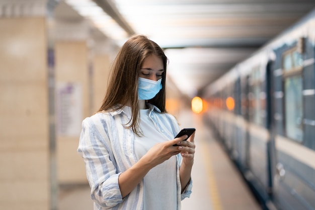 Vrouw draagt gezichtsmasker met behulp van mobiel chatten op sociale media, wachtend op metro op perron