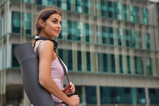 Vrouw draagt yogamat gaat oefensessie doen gekleed in sportkleding luistert muziek via koptelefoon poseert buitenshuis