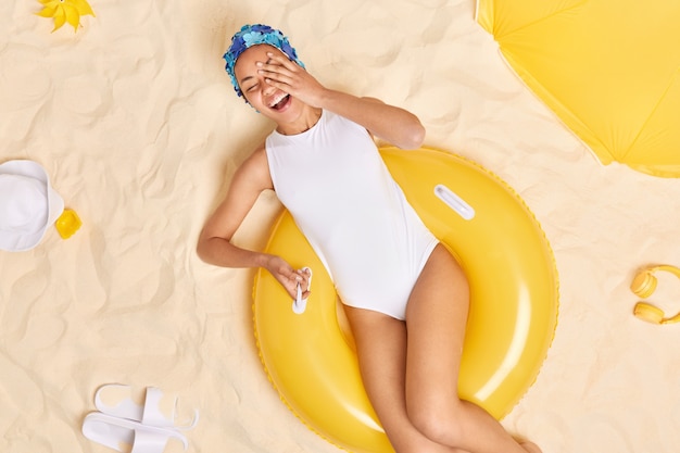 Vrouw draagt zwemhoed en witte bikini poseert op opgeblazen zwemband brengt vrije tijd door aan zandstrand gele parasol koptelefoon panama en slippers rond