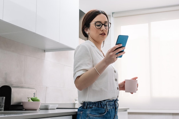 Vrouw drinkt koffie in de keuken terwijl ze haar smartphone gebruikt