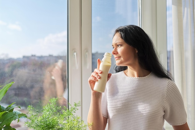 Vrouw drinkt melkdrank uit een fles die bij het raam staat melk yoghurt zuivel gezonde dranken