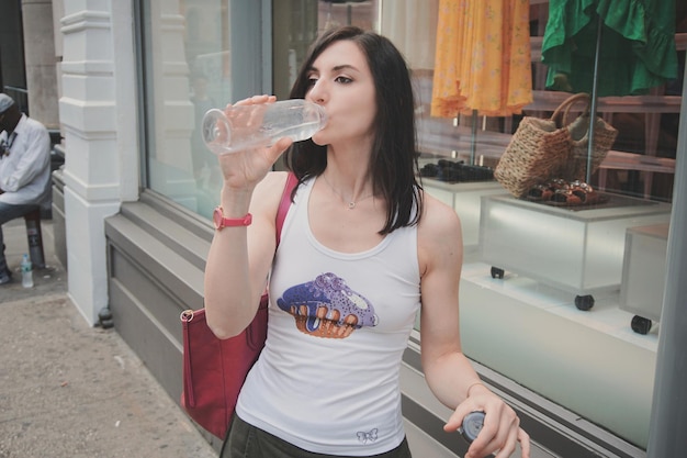 Foto vrouw drinkt water terwijl ze op het voetpad staat tegenover een winkel in de stad