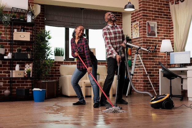 Vrouw dweilt de vloer en danst terwijl de man met passie zingt met een stofzuiger. Afro-Amerikaans echtpaar dat in het weekend het huis schoonmaakt en ervoor zorgt dat het huis netjes en schoon blijft.