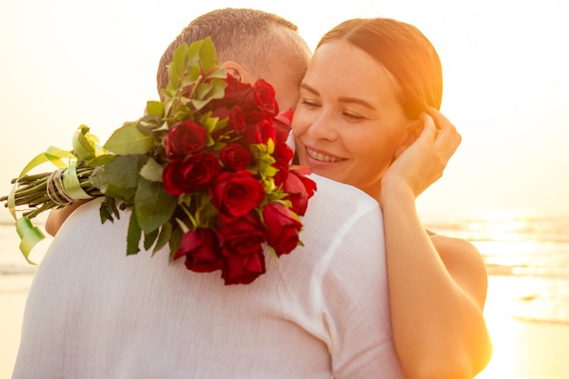 Vrouw en man gelukkig paar op vakantie aan zee mooie vrouw in rode jurk met een ring aan haar vinger en liefdevolle man in een wit overhemd met een boeket rozen romantische zonsondergang Valentijnsdag datum