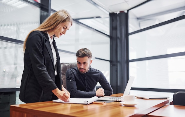 Vrouw en man in formele kleding werken binnen op kantoor samen aan tafel.