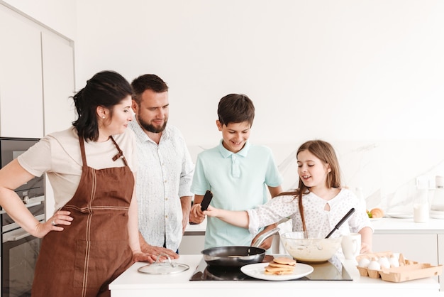 Vrouw en man met gelukkige kinderen 8-10 samen koken en pannenkoeken bakken op modern fornuis in keuken thuis