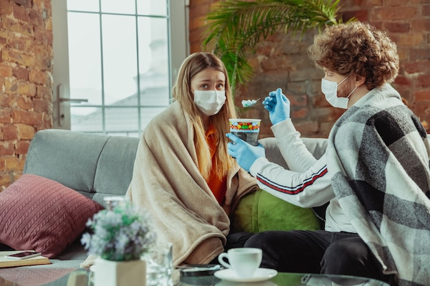 Vrouw en man, paar met beschermende maskers en handschoenen thuis geïsoleerd met luchtwegsymptomen van het coronavirus zoals koorts, hoofdpijn, hoest. gezondheidszorg, medicijnen, quarantaine, behandelingsconcept.