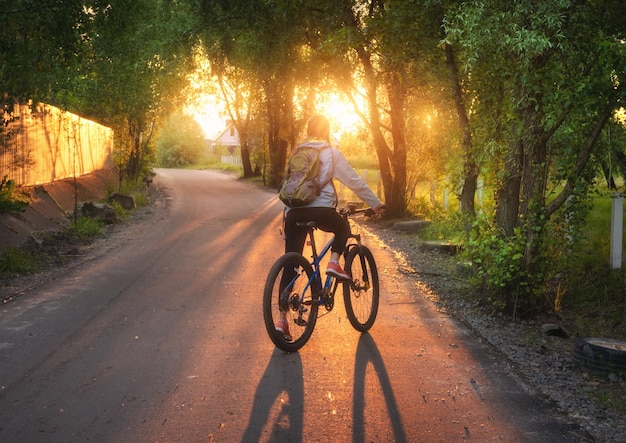 Vrouw fietsen op de landelijke weg bij zonsondergang in de zomer Kleurrijk landschap met sportief meisje met rugzak rijden op een mountainbike weg groene bomen en goud zonlicht Sport en reizen cyclus