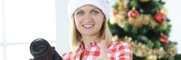 Vrouw fotograaf in kerstman hoed op de achtergrond van de kerstboom maakt duimen omhoog gebaar