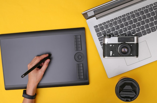 Vrouw fotograaf of retoucher werken met grafisch tablet, camera en laptop op gele achtergrond