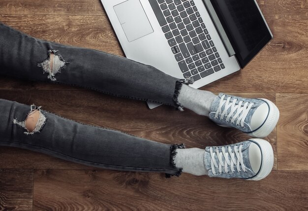 Vrouw freelancer in gescheurde spijkerbroek en sneakers zittend op een houten vloer met laptop.