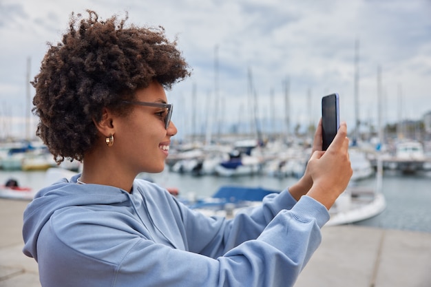 vrouw gaat op cruiseschip vakantie geniet van uitzicht op zeehaven maakt selfie op moderne smartphone draagt zonnebril en hoodie neemt foto van zichzelf in haven