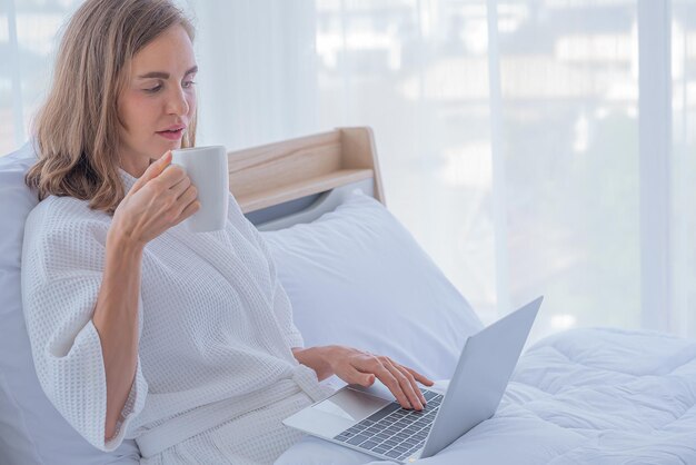 Foto vrouw gebruikt computernotitieboekje op bed en drinkt koffie