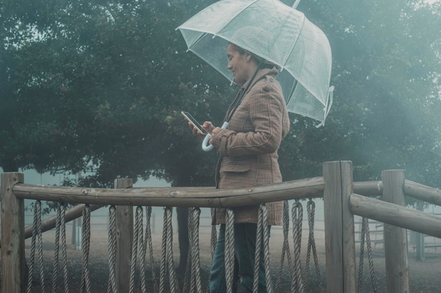 Vrouw gebruikt mobiele telefoon onder de regen beschermd tegen een transparante paraplu Regenachtige dag en slechte weersomstandigheden in het park Mensen in openluchtrecreatie in de herfst of winter Dame met Britse stijl