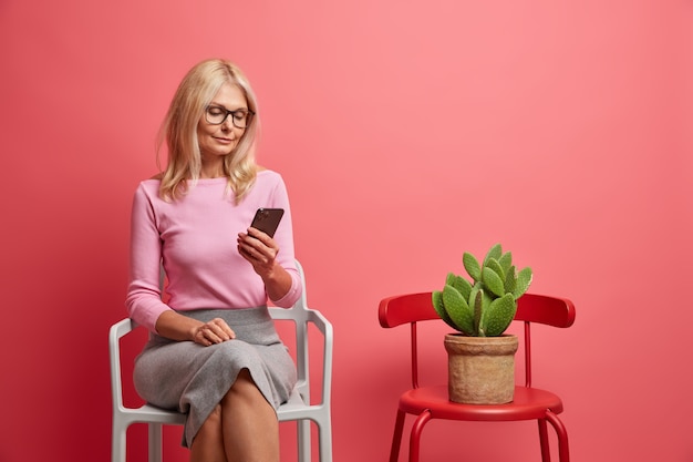 vrouw geconcentreerd op smartphone leest nieuws online draagt nette kleding poses in de buurt van stoel met cactus in pot geïsoleerd op roze