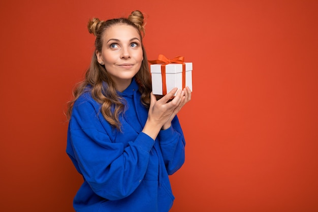 Vrouw geïsoleerd op kleurrijke achtergrond muur dragen stijlvolle casual kleding bedrijf geschenkdoos en op zoek naar de zijkant. kopieer ruimte