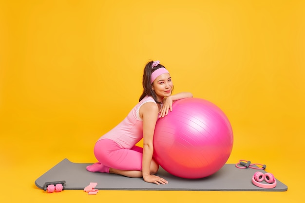 vrouw gekleed in bodysuit poseert op yogamat met fitnessbal maakt gebruik van elastische tape en verschillende sportuitrusting geïsoleerd op geel