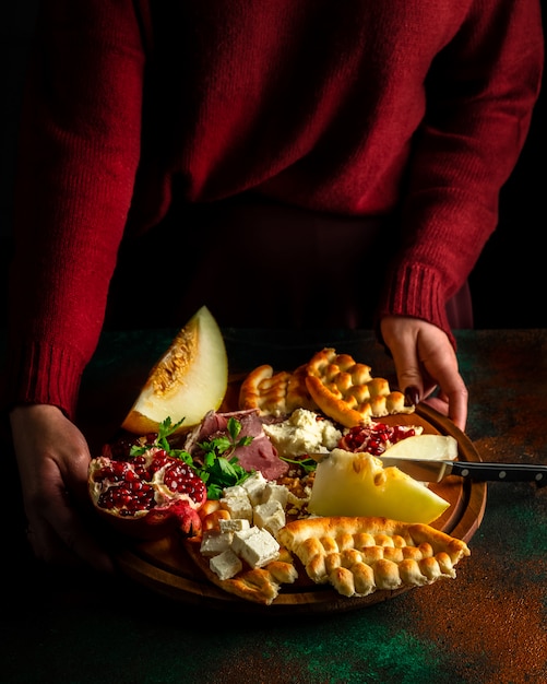 Vrouw gekleed in donkerrode trui met delicatessen bord met gerookte ham, witte kaas, fruit en plat brood