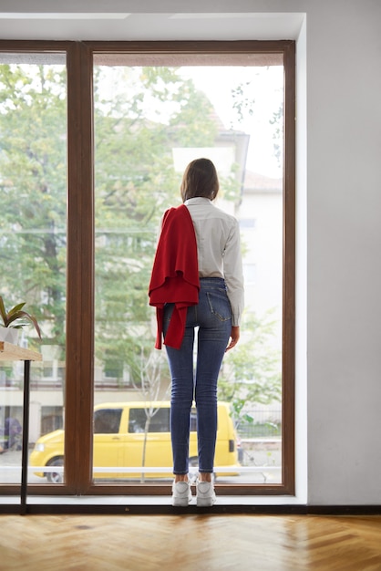 Foto vrouw, gekleed in een wit overhemd en een spijkerbroek staan in de buurt van het raam