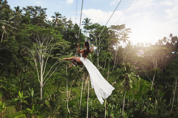 Vrouw, gekleed in een witte jurk, slingert op touwschommels met prachtig uitzicht op rijstterrassen en palmbomen op het eiland Bali