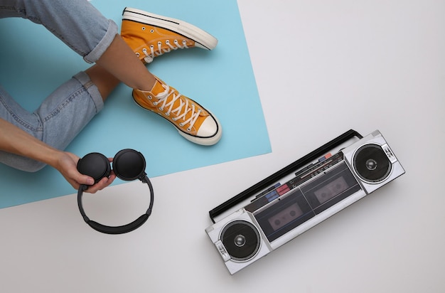 Vrouw gekleed in spijkerbroek en sneakersh met stereo koptelefoon, retro audio cassetterecorder op een blauw-witte achtergrond. Retro jaren 80 cultuur. Bovenaanzicht. plat leggen