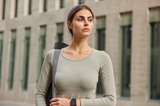 vrouw gekleed in sportkleding draagt karemat op schouder draagt smartwatch kijkt in afstand staat in de buurt van gebouw rust na lichamelijke activiteit