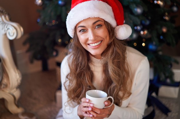 Vrouw gekleed witte trui kerstmuts en spijkerbroek zittend op de vloer in de buurt van kerstboom met huidige doos