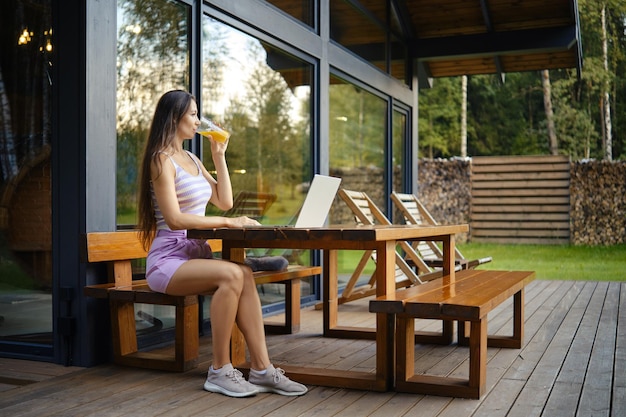 Vrouw geniet van een drankje terwijl ze een laptop gebruikt op het patio van een moderne hut omringd door de natuur