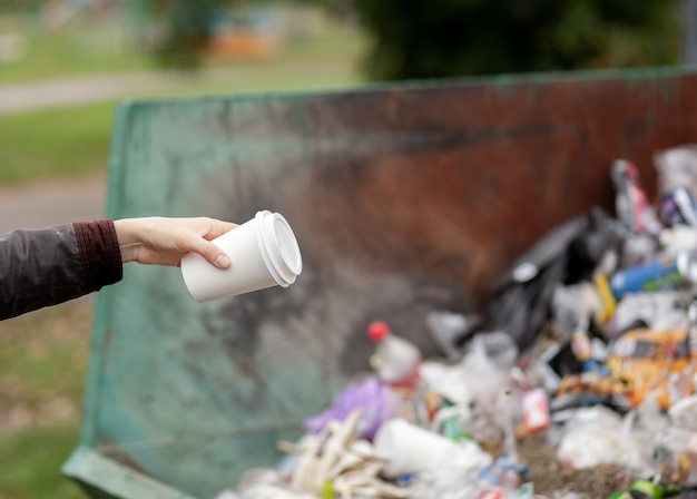 Vrouw gooit een kartonnen glas in een prullenbak. Zorg dragen voor de netheid van de stad en het milieu. Een grote vuilnisbak in een openbaar park