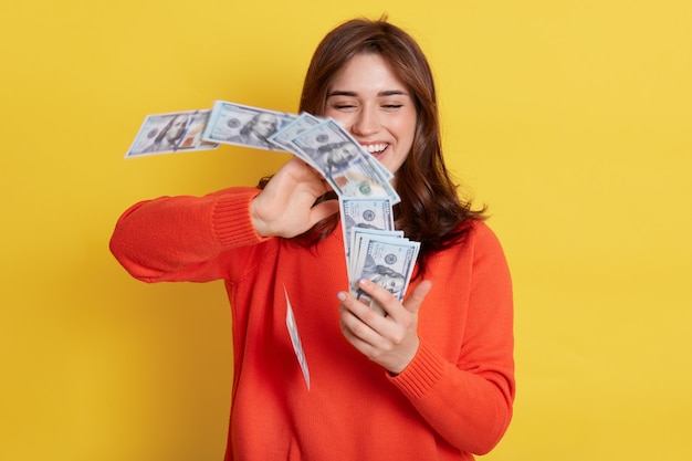 Vrouw gooit in luchtgeld, lacht vrolijk, houdt een grote som geld vast, heeft beloning voor werk, draagt een oranje trui, een rijke dame die een loterij wint, poseren met valuta geïsoleerd over een gele muur.