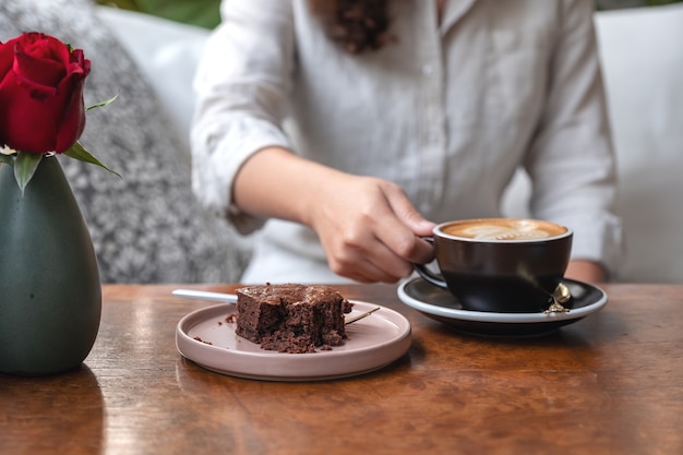 Vrouw hand houden en drinken van hete latte koffie met brownie cake op tafel