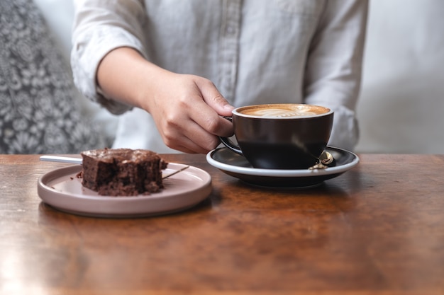 Vrouw hand houden en drinken van hete latte koffie met brownie cake op tafel