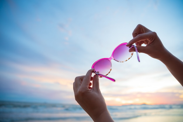 Vrouw hand met zonnebril over zee en zandstrand tijdens zonsondergang