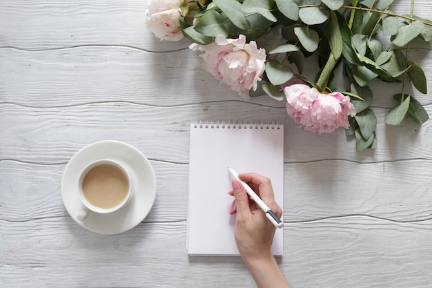 Vrouw hand schrijft in een notitieblok. Tafel met pioenrozen en een kopje koffie bovenaanzicht.