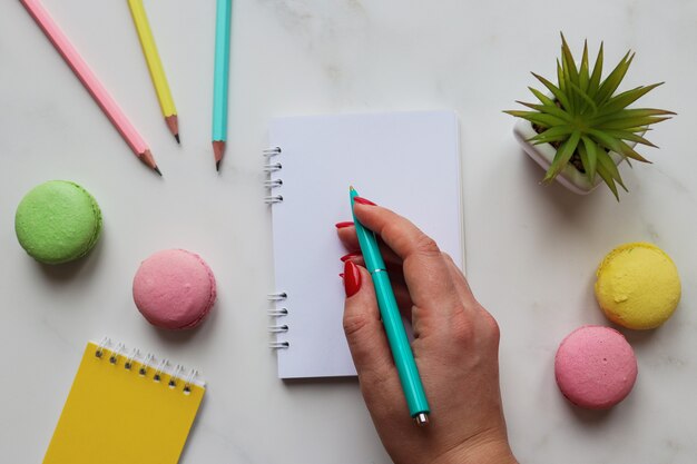 Vrouw hand schrijven in notitieblok. werkruimte met notitieboekjes, potloden, pen, plant en bitterkoekjes