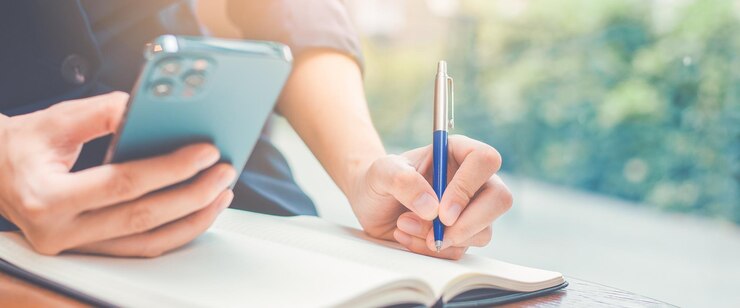 Vrouw hand schrijven op een notitieblok met een pen en ze houdt een mobiele telefoon in de office.Web banner.