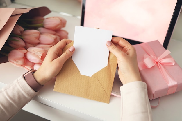 Vrouw handen met een brief in ambachtelijke envelop. Roze achtergrond, Valentijnsdag concept. Tulpenbloem en roze geschenkdoos op de achtergrond. Thuis bureau voor dames.