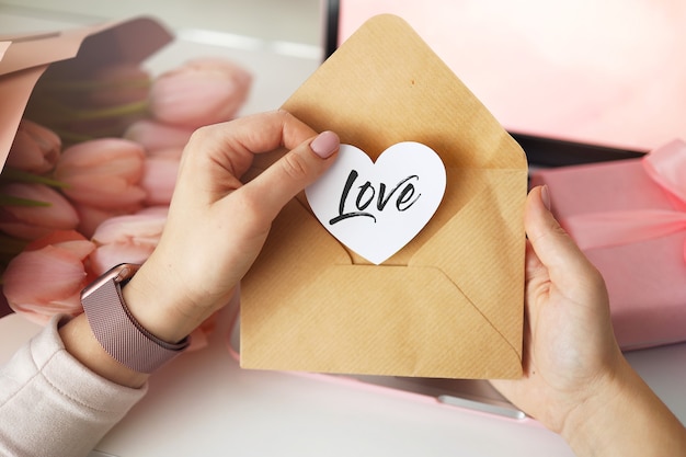 Foto vrouw handen met een brief in ambachtelijke envelop. roze achtergrond, valentijnsdag concept. tulpenbloem en roze geschenkdoos op de achtergrond. thuis bureau voor dames.