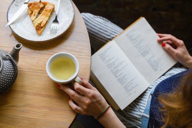 Vrouw handen met geopend boek kopje thee taart op lichte houten tafel