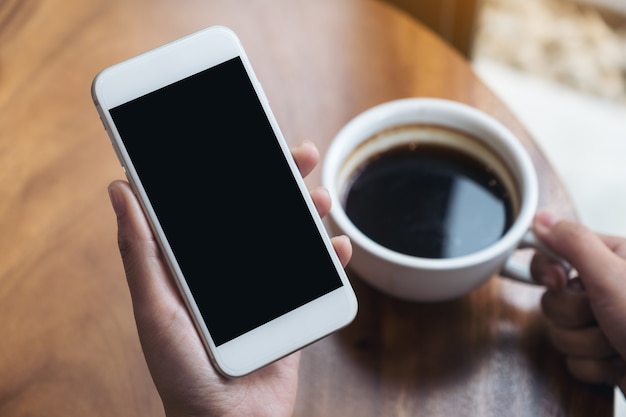 vrouw handen met witte mobiele telefoon met leeg zwart scherm terwijl het drinken van koffie