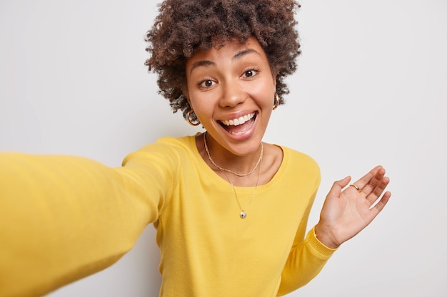 vrouw heeft blije gezichtsuitdrukking strekt arm uit om selfie te maken drukt oprechte emoties uit gekleed in casual gele trui poseert op wit neemt selfie