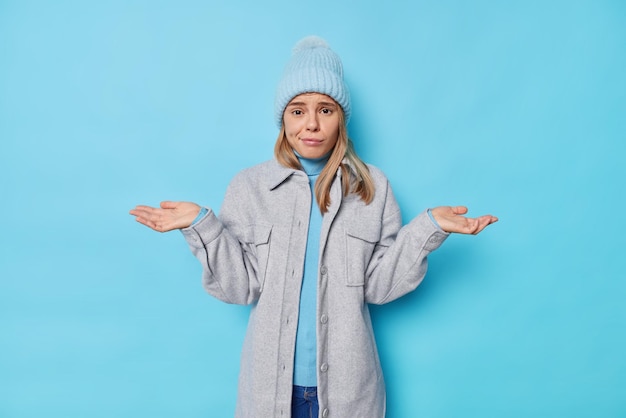 Foto vrouw heeft vragen drukt teleurstelling uit haalt haar schouders op met handen voelt besluiteloos voelt aarzelend draagt hoed en jas geïsoleerd op blauw weet niet wat te doen.