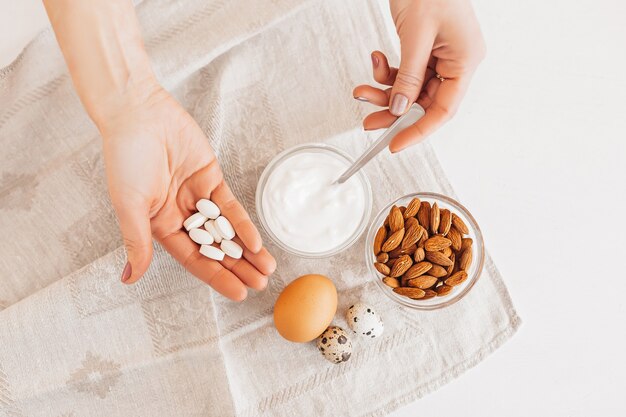 Vrouw houdt een handvol voedingssupplementenpillen vast. Noten amandelen, zure room of yoghurt, kip en kwarteleitjes op een linnen keukenhanddoek op een witte tafel. Gezonde voeding, voedingsstoffen vitaminen concept