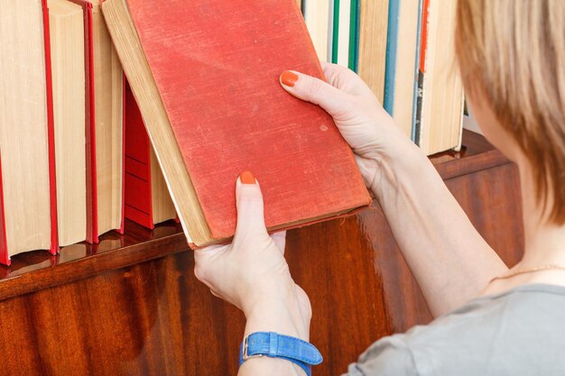 Vrouw houdt een oud boek naast een boekenplank. Veel boeken met harde kaft op houten plank. Bibliotheekconcept