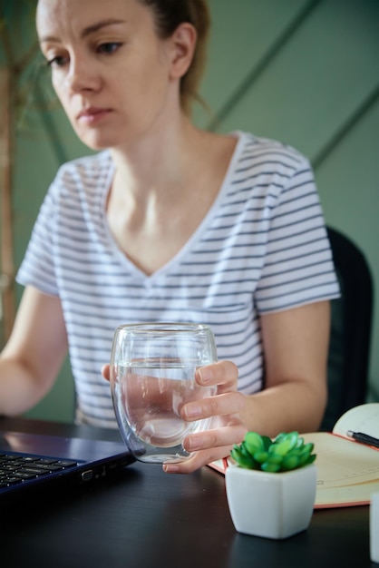 Vrouw houdt glas water vast terwijl ze op afstand werkt op kantoor aan huis gezonde zorg voor freelancer werk